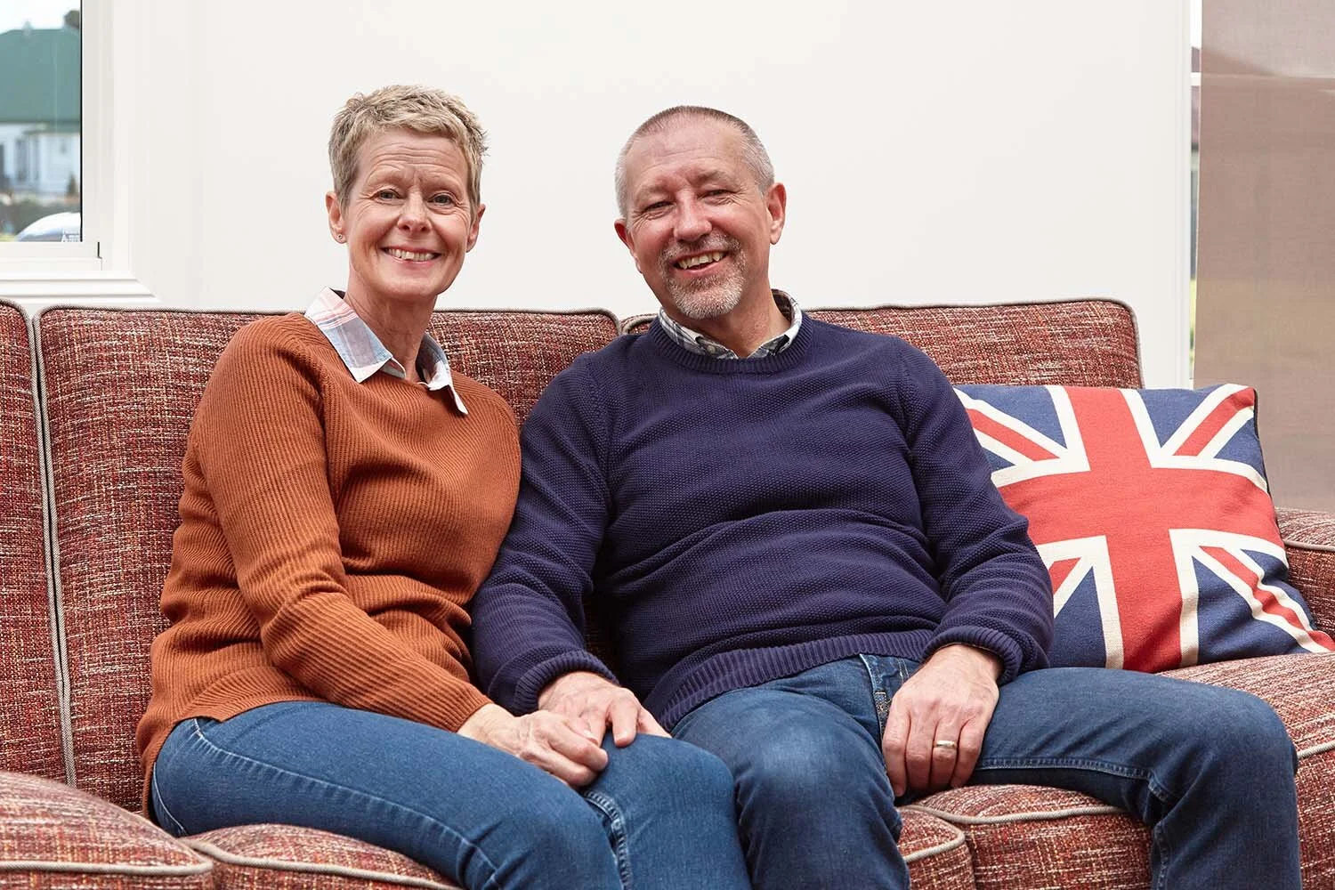 Smiling older couple sitting on a sofa with a United Kingdom flag pillow.