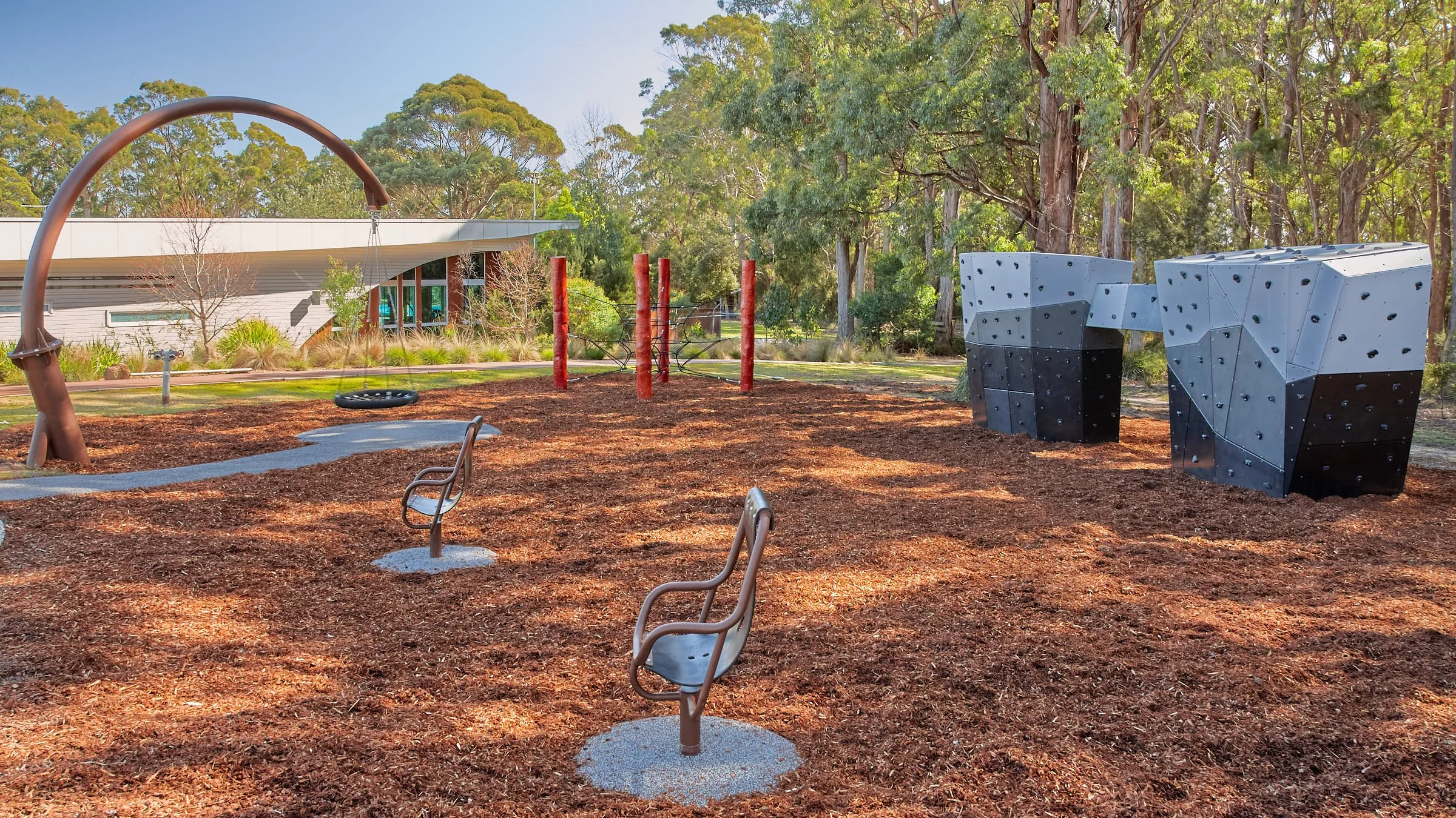 Children's outdoor playground with spinning tire, climbing structures, and benches, surrounded by trees and mulch.