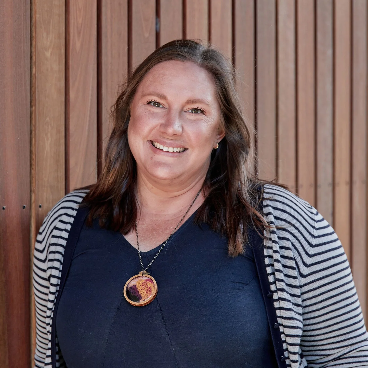 Smiling woman with long brown hair, wearing a navy blue top, a striped cardigan, and a colorful pendant necklace, standing in front of a wooden wall.