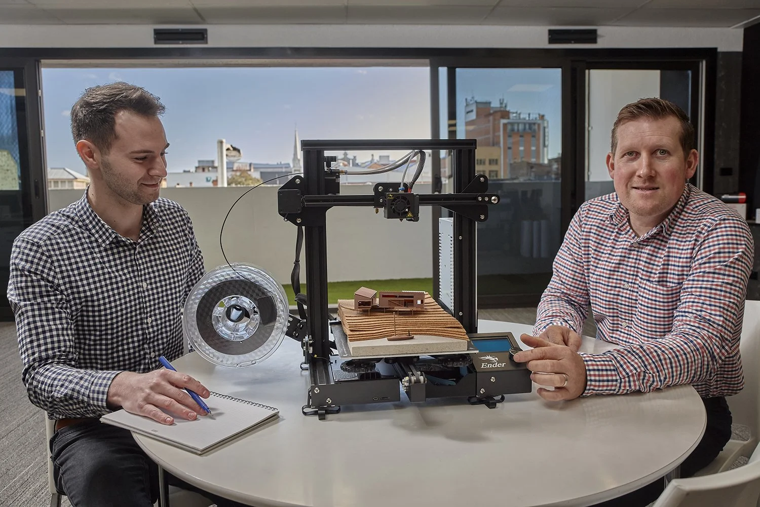 Two men in checkered shirts seated at a table with a 3D printer and architectural models, in a room with large windows and city buildings outside.