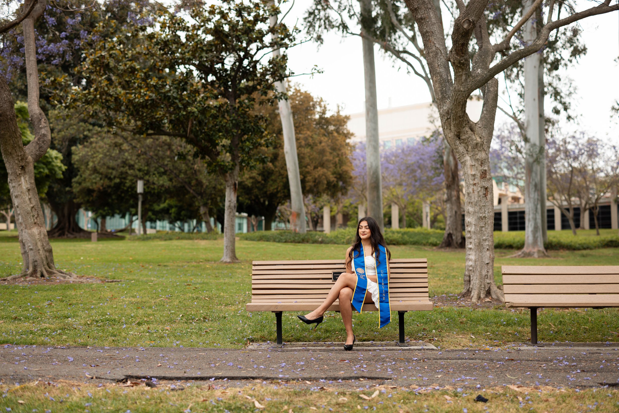 A young woman in a white dress with a blue graduation stole sitting on a park bench, smiling, with trees and purple flowers in the background.