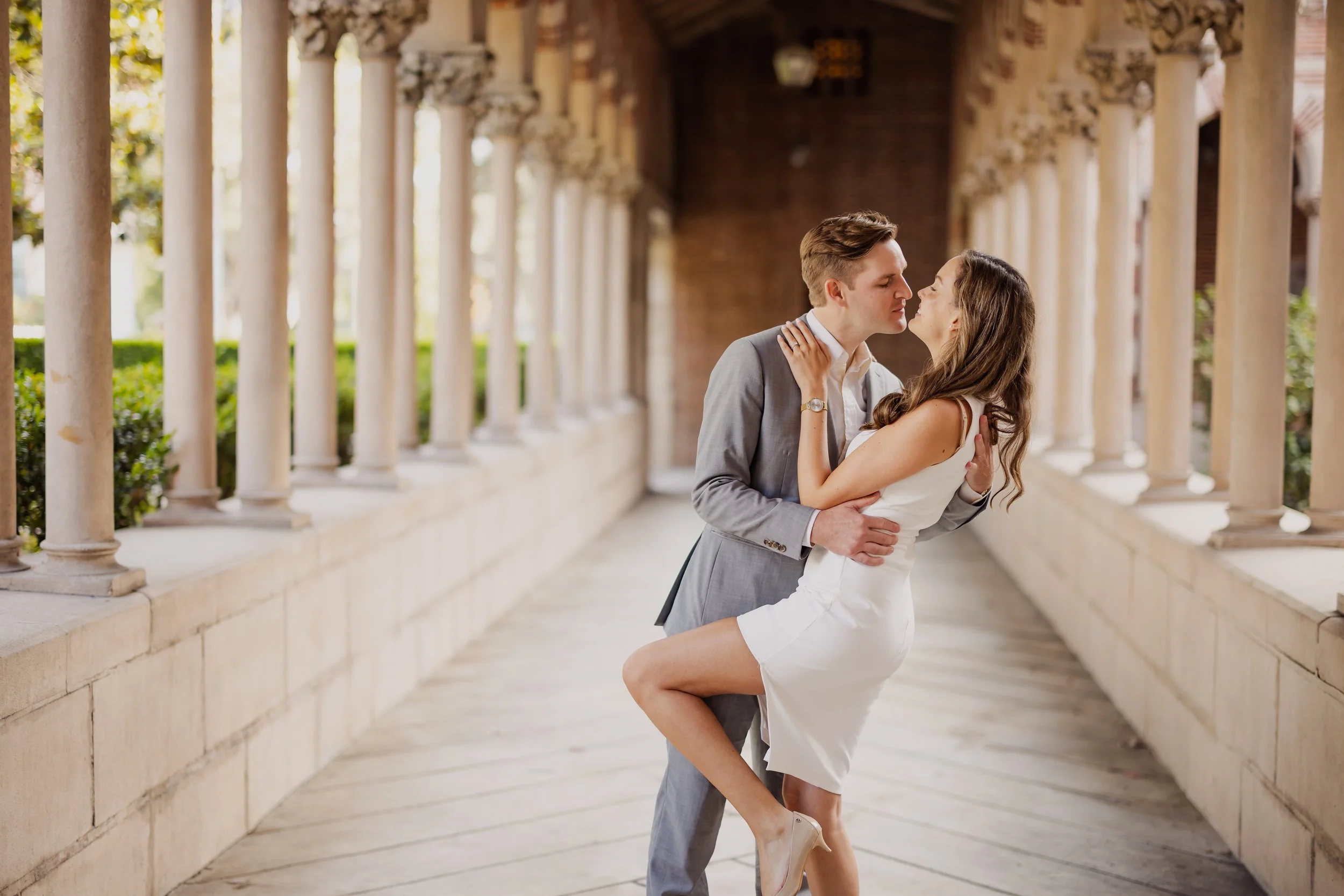 A couple in formal attire sharing an intimate moment in a scenic outdoor walkway with stone columns and greenery in the background.