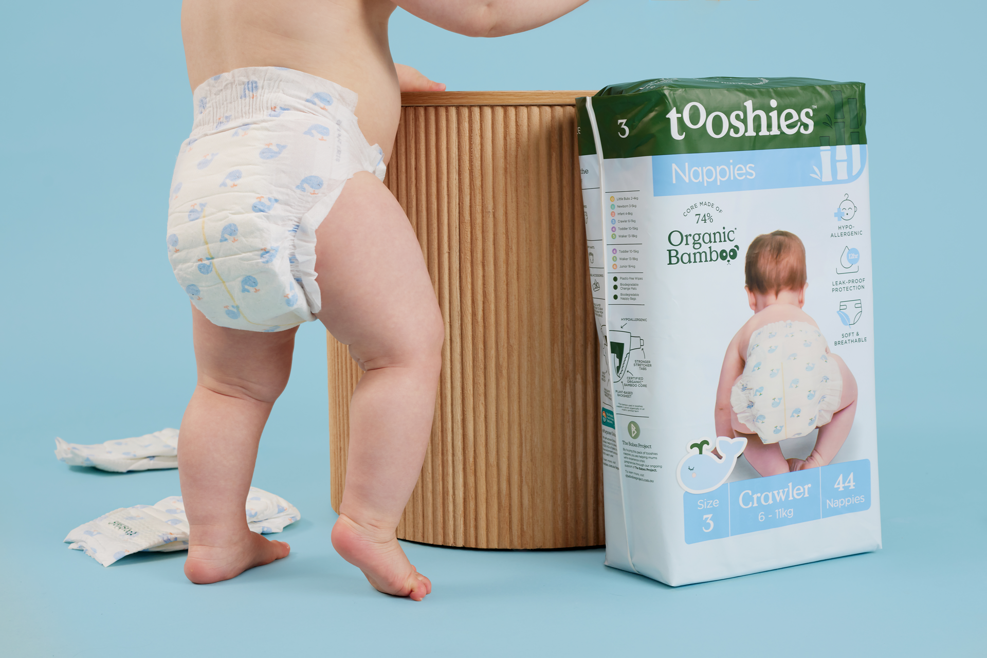 A baby crawling on a beige surface, wearing a diaper with purple flowers and green leaves, with a light-colored blanket in the background.