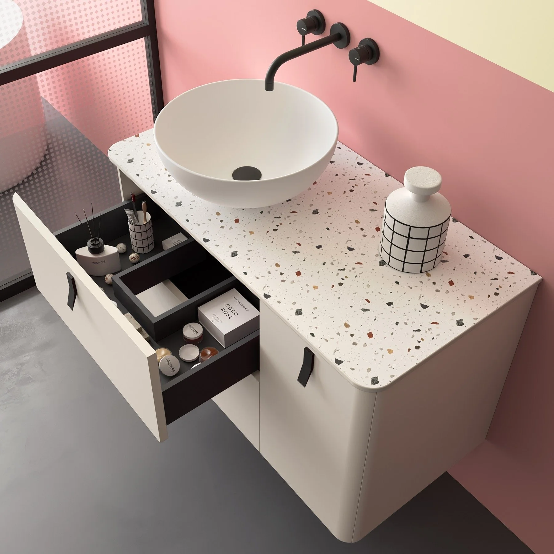 Modern bathroom vanity with a round white terrazzo sink, black faucet and knobs, pink wall panel, a container with a grid pattern, and drawers with small bathroom products inside. Modern bathroom furniture available from Island Bathrooms in Dorset