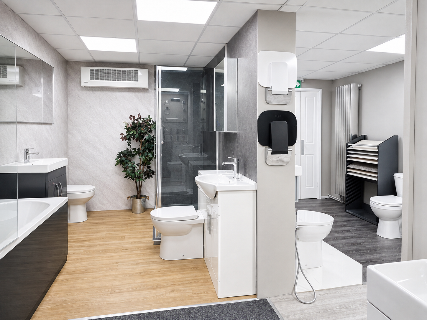 A modern bathroom with a dual-level layout; the front area has a black vanity and a toilet, the back area features a shower, a toilet, and a sink, with various paper towel dispensers and a potted plant.