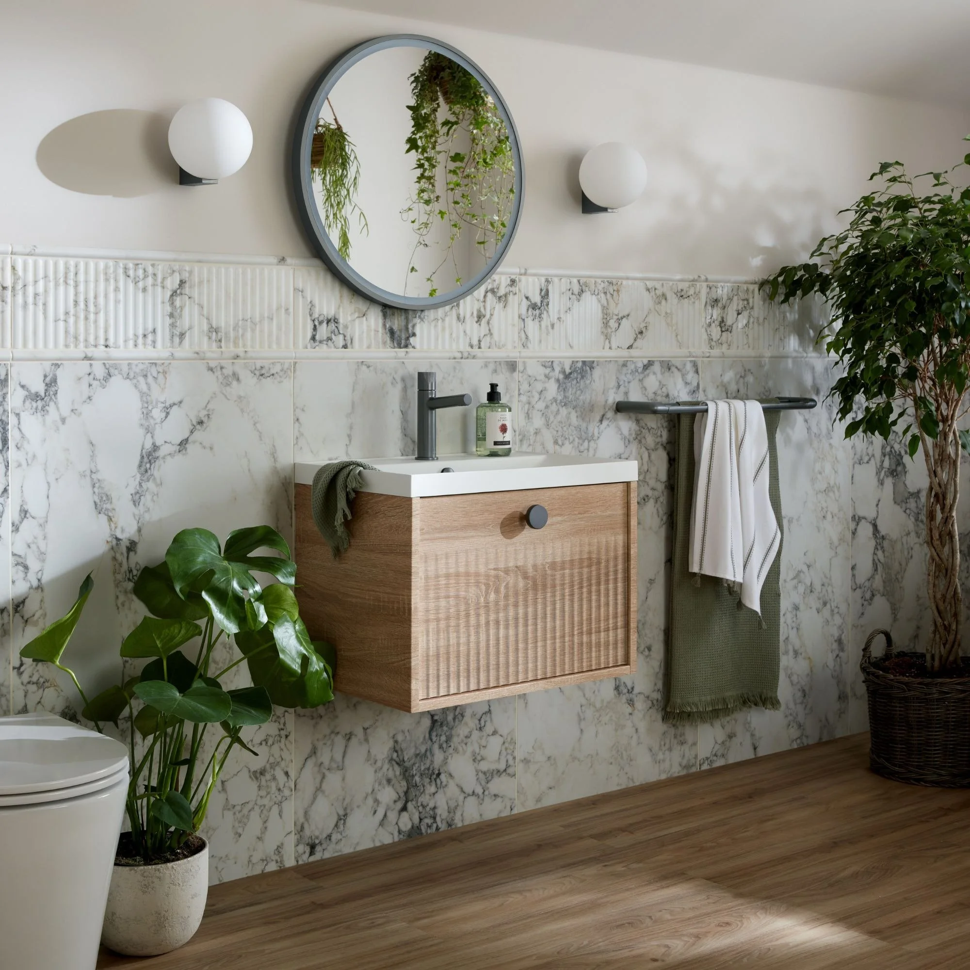 Modern bathroom with a round mirror, two wall-mounted white globe lights, a small wooden vanity with a white sink, green plants in white pots, marble backsplash, and wood flooring.