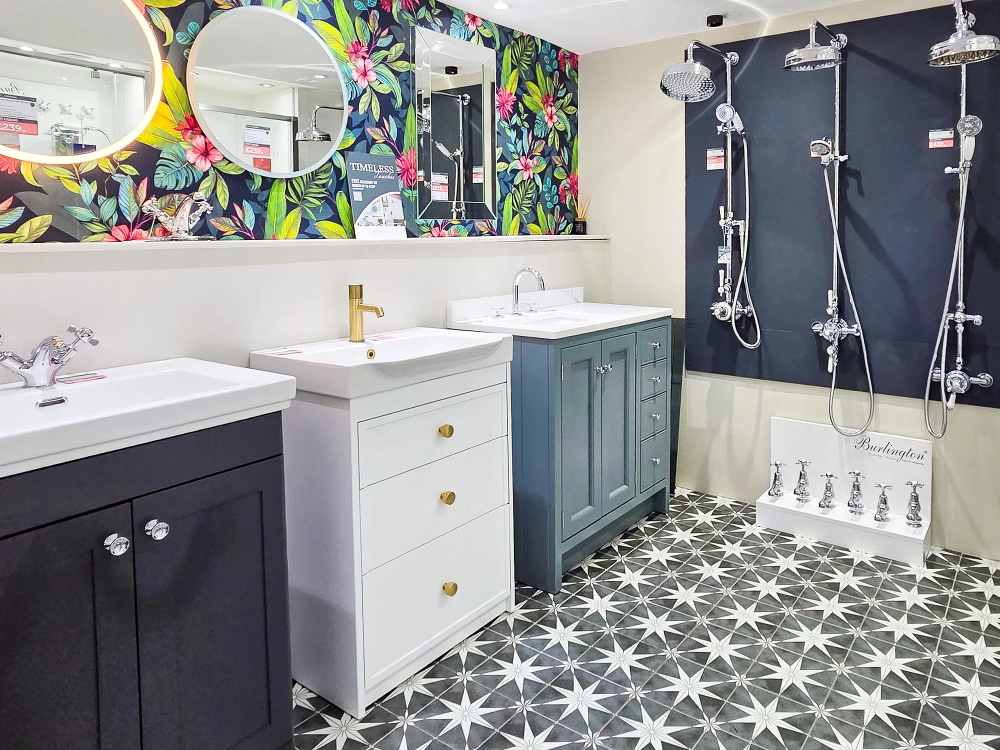 Display of bathroom fixtures including several shower heads mounted on a dark wall, with decorative floral wallpaper and a large mirror, along with two sinks, and a floor with black and white star-patterned tiles.
