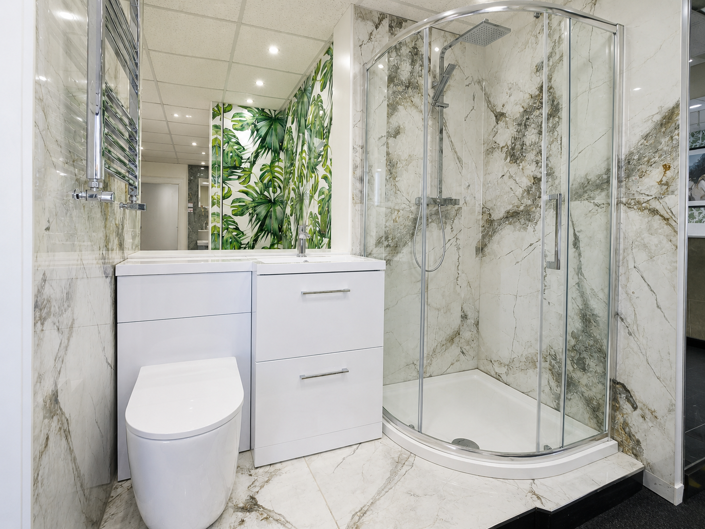 Modern bathroom with marble walls and floors, a white toilet, a white vanity with drawers, a mirrored cabinet, and a curved glass shower enclosure. There is green leafy wallpaper or mural behind the sink area.