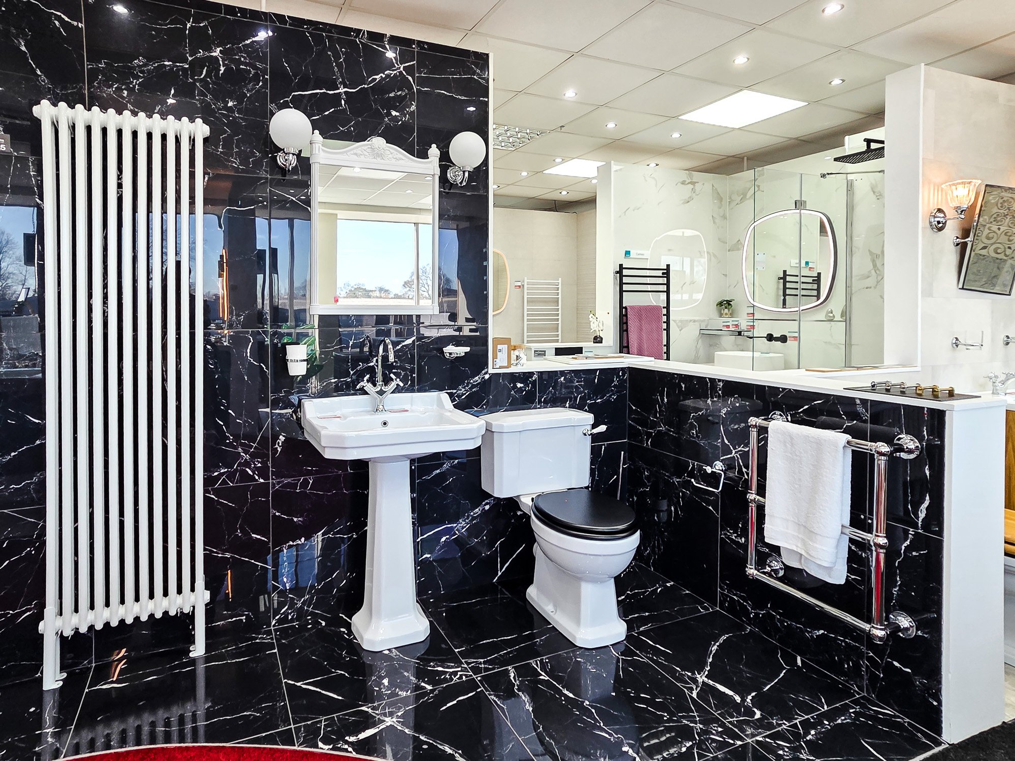 A bathroom with black marble tiles on the wall and floor, featuring a white sink, a mirror, a heated towel rack, and a divided shower area with glass panels. There are round light fixtures and a window in the background.