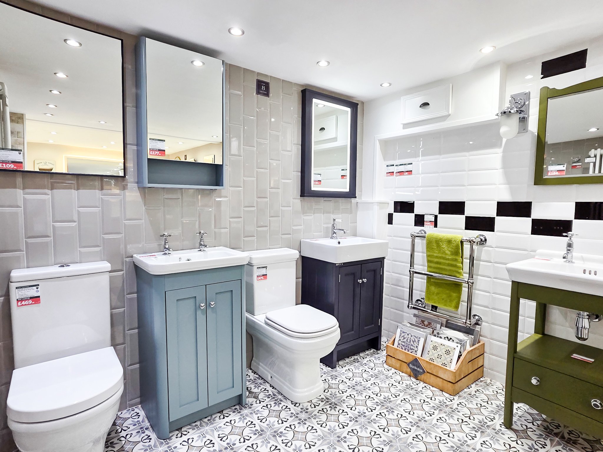 A display of bathroom sinks, toilets, and mirrors in a home improvement store, with patterned floor tiles and various price tags.