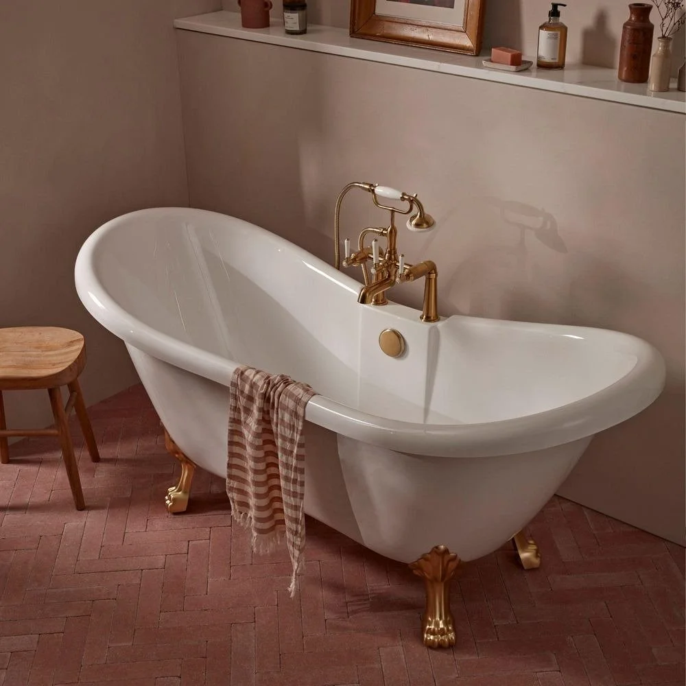 A vintage clawfoot Burlington bathtub with gold fixtures in a bathroom, with a striped towel hanging over the side and a small wooden stool nearby. Traditional bathroom design available from Island Bathrooms Bournemouth, Dorset