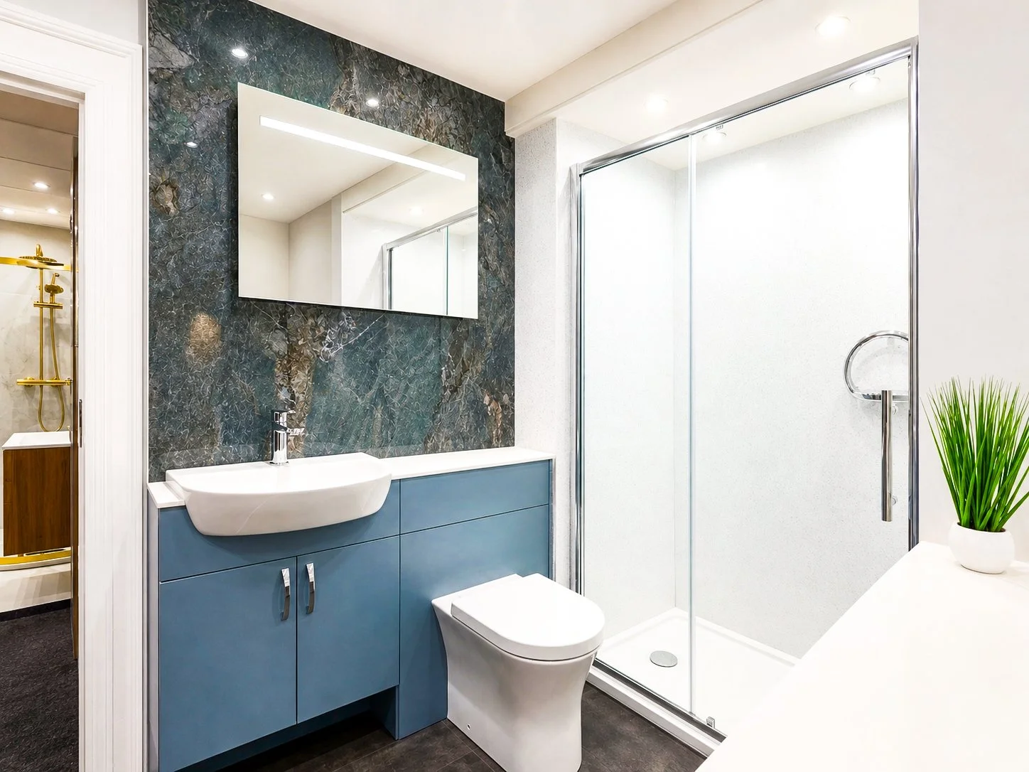 Modern bathroom with a small sink, blue cabinet, mirror, transparent shower enclosure, a potted plant, and gold fixtures in the shower area.