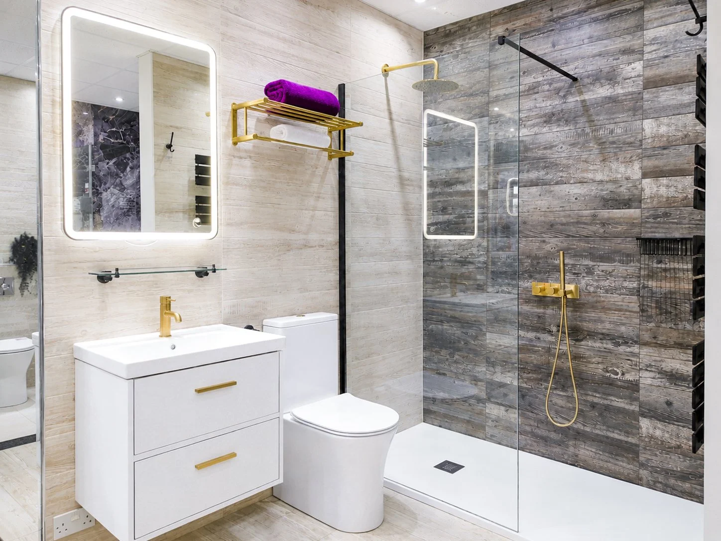 Modern bathroom with a white vanity, gold fixtures, a toilet, and a walk-in shower with dark wood tiles and gold fixtures, illuminated mirror, and wall-mounted towel rack with purple and white towels.