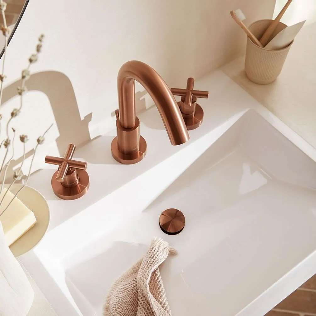 Close-up of a modern bathroom sink with a matte copper faucet and matching handles, a light beige soap dish, and a container with toothbrushes and a wooden spoon. A hand towel hangs over the sink edge.