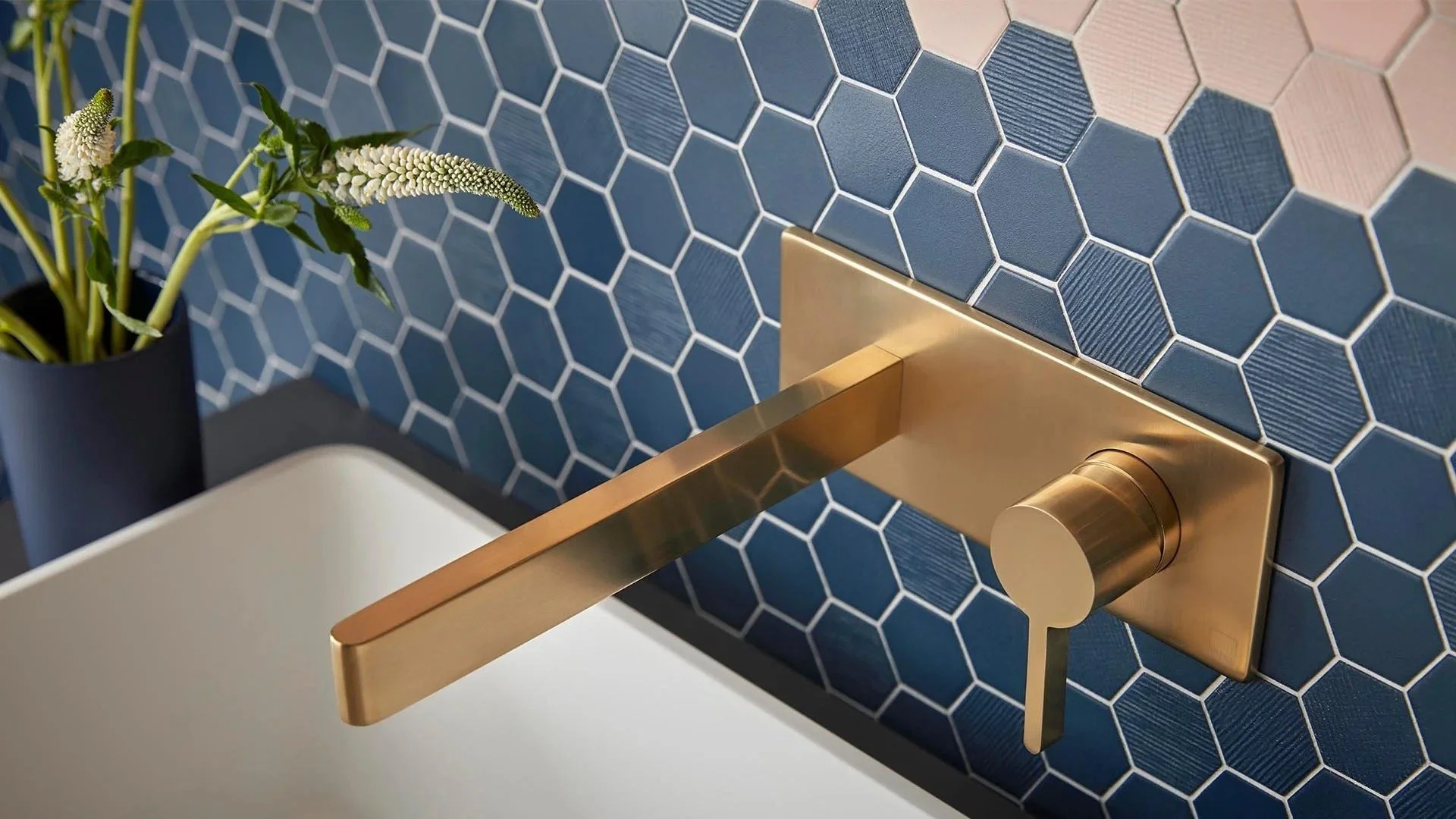Close-up of a modern gold door handle mounted on a gold plate, against a hexagonal tile wall in shades of blue, with a black vase holding green and white flowers in the background. bathroom taps available at Island Bathrooms Bournemouth and Salisbury
