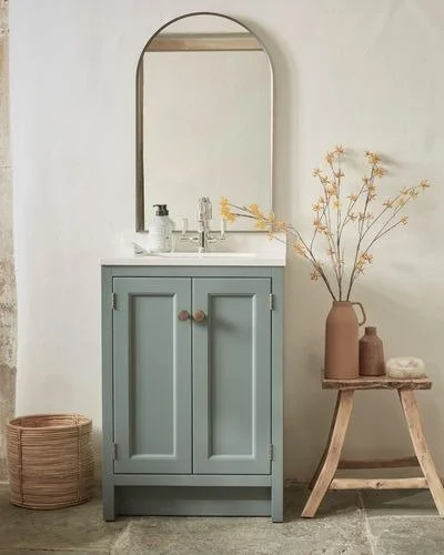 A bathroom vanity with a light blue cabinet, a white countertop, and a mirror above. Next to it, there is a wooden side table with vases and dried flowers, and a woven basket on the floor.