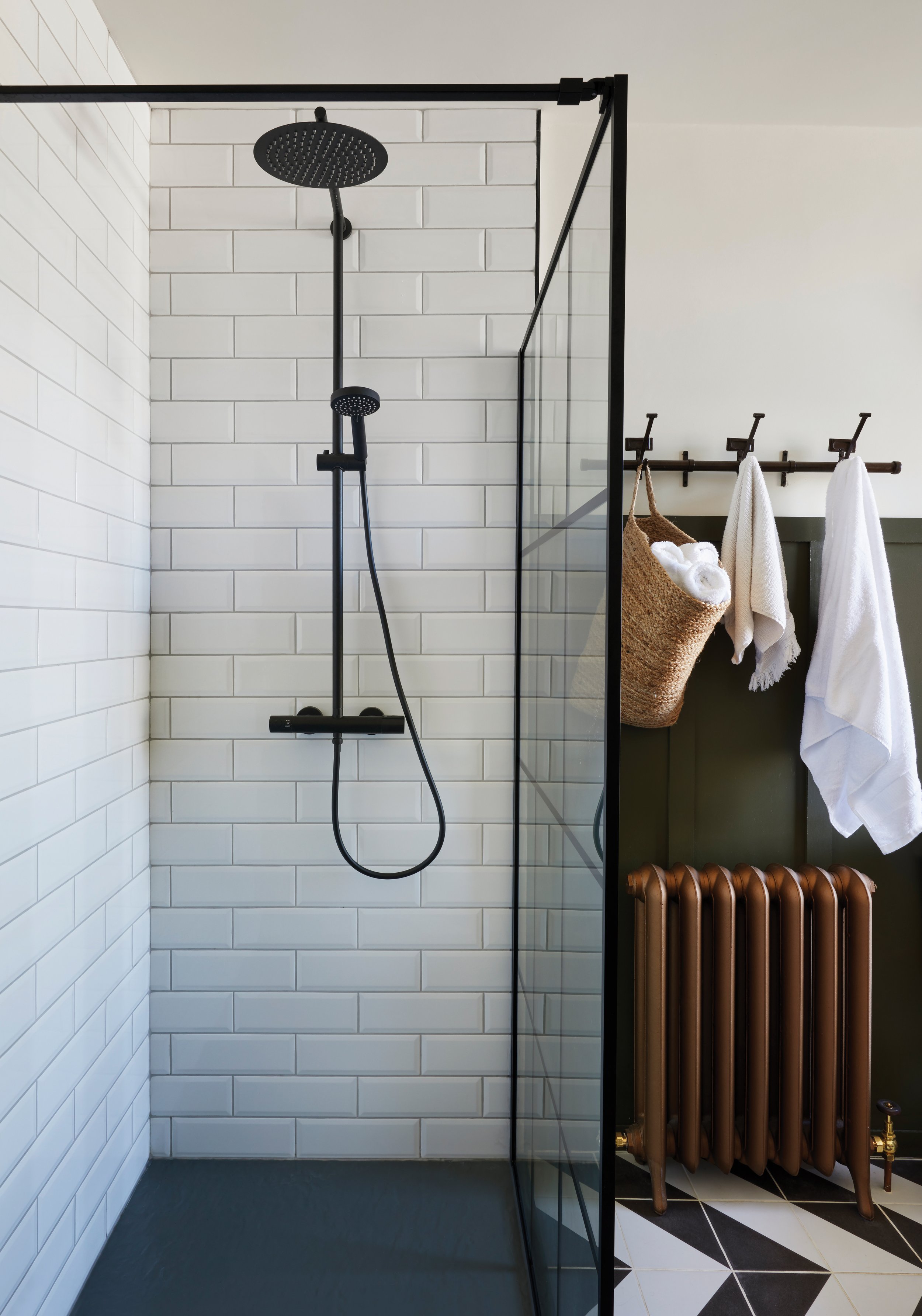 Modern walk-in shower with black fixtures and white subway tile walls, adjacent to a dark green wall with a brown radiator, a wall-mounted towel rack with white towels, a hanging wicker basket with rolled towels, and black-and-white patterned floor tiles.