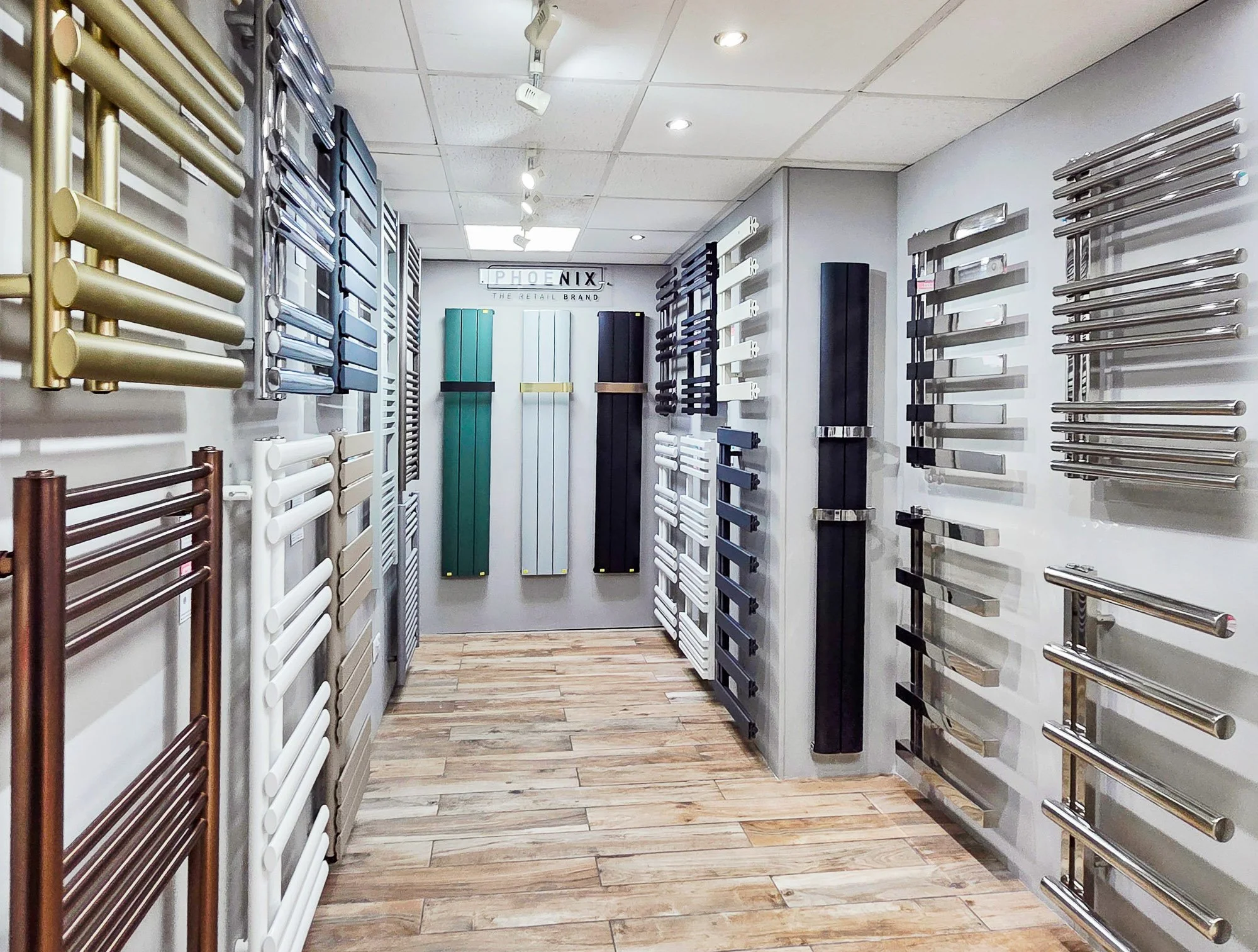 Display of various towel radiators and heated towel rails in a showroom. The walls are lined with different styles and colors of towel radiators, including brass, chrome, black, white, and colored options. The shop has a wooden floor and a ceiling wi