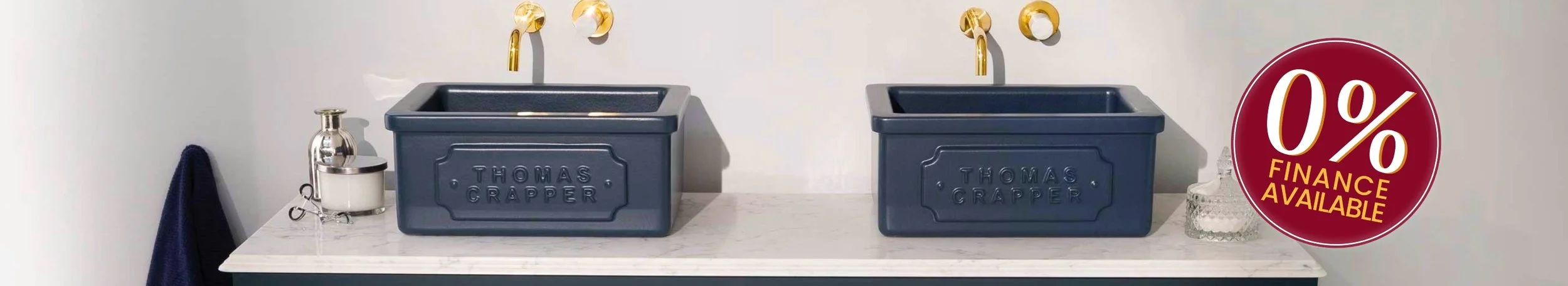 Two dark gray laundry sinks labeled 'Thomas Crapper' installed side by side on a marble countertop with a small glass jar and a soap dispenser nearby. A navy towel hangs on a hook on the left, and a red circular sign indicating '0% finance available'