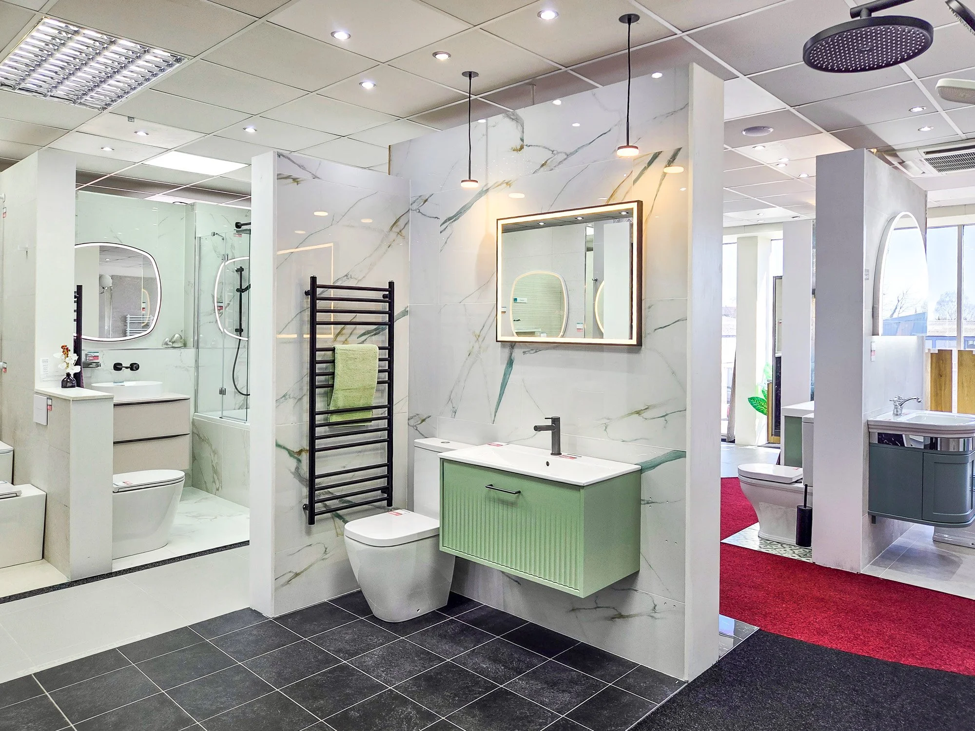 A modern bathroom display in a showroom with marble walls, a wall-mounted toilet, a green sink vanity, and mirrors. The showroom has ceiling lights, a heated towel rack, and visible natural light from large windows.