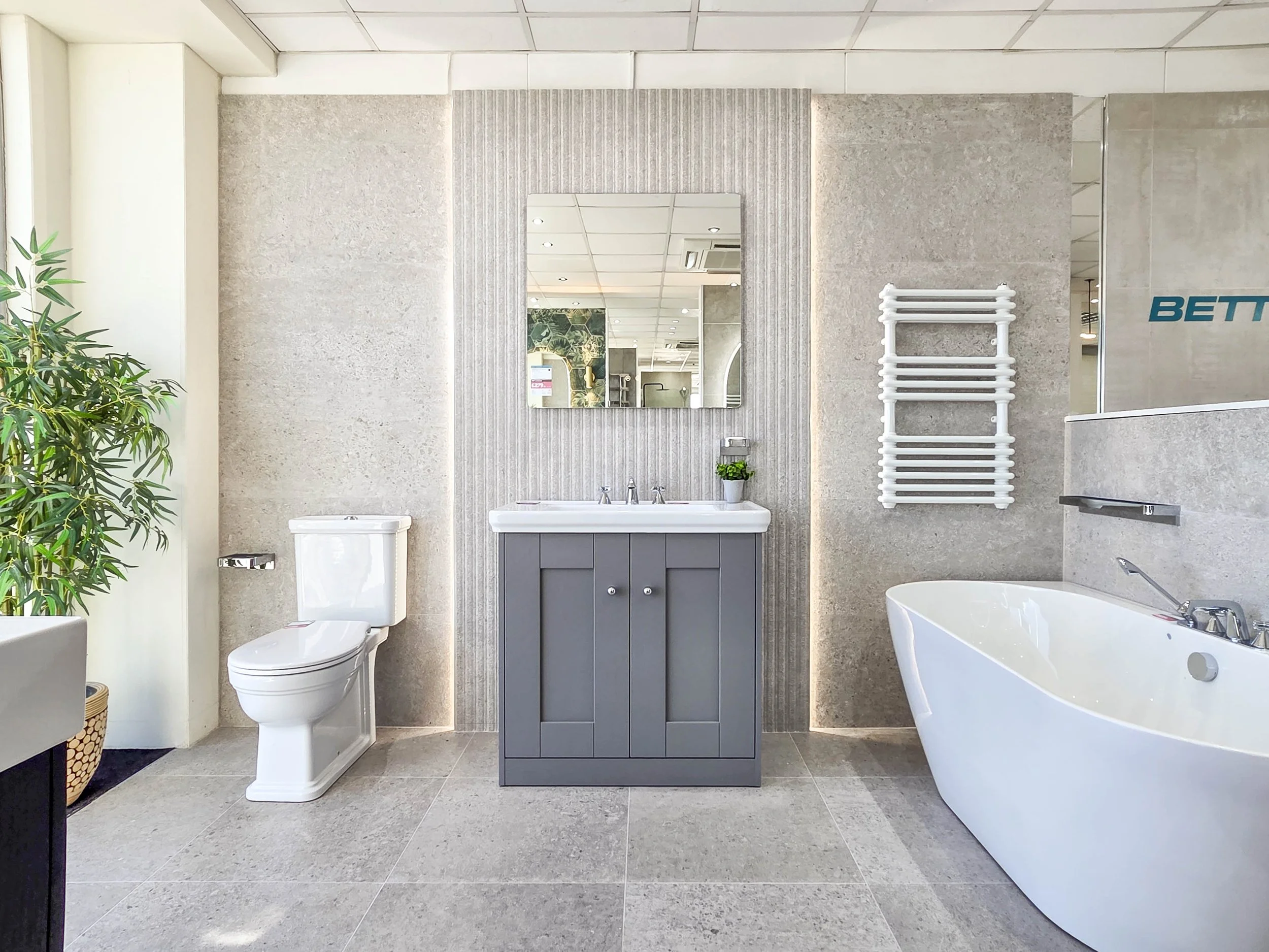 A modern bathroom with a bathtub, toilet, sink with mirror, towel radiator, and potted plant, with light-colored tiled walls and floor.