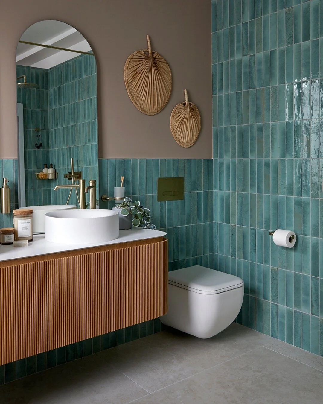 Modern bathroom with teal tiled walls, a wooden vanity with a white vessel sink, and a wall-mounted toilet. Decor includes two dried palm leaf wall hangings and some green plants, with a large mirror reflecting part of the shower area.