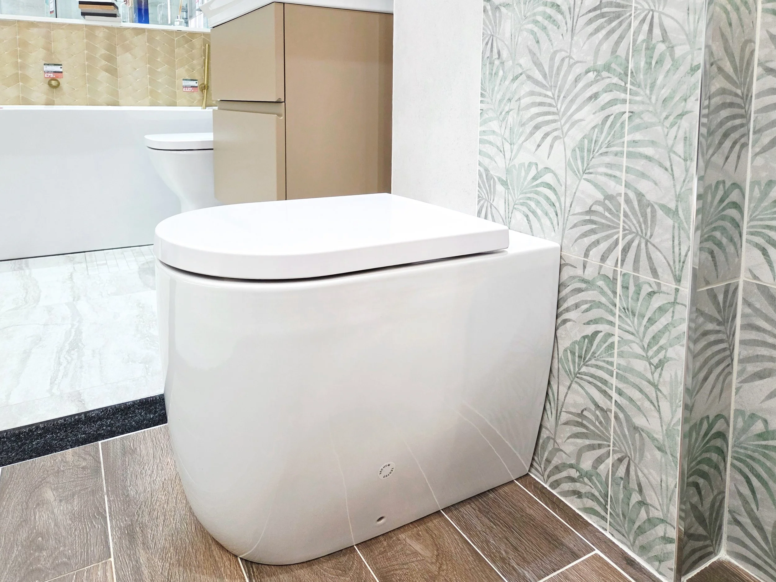 A modern, white bidet toilet in a bathroom with leafy patterned wall tiles, wood-look floor tiles, and a beige cabinet in the background.