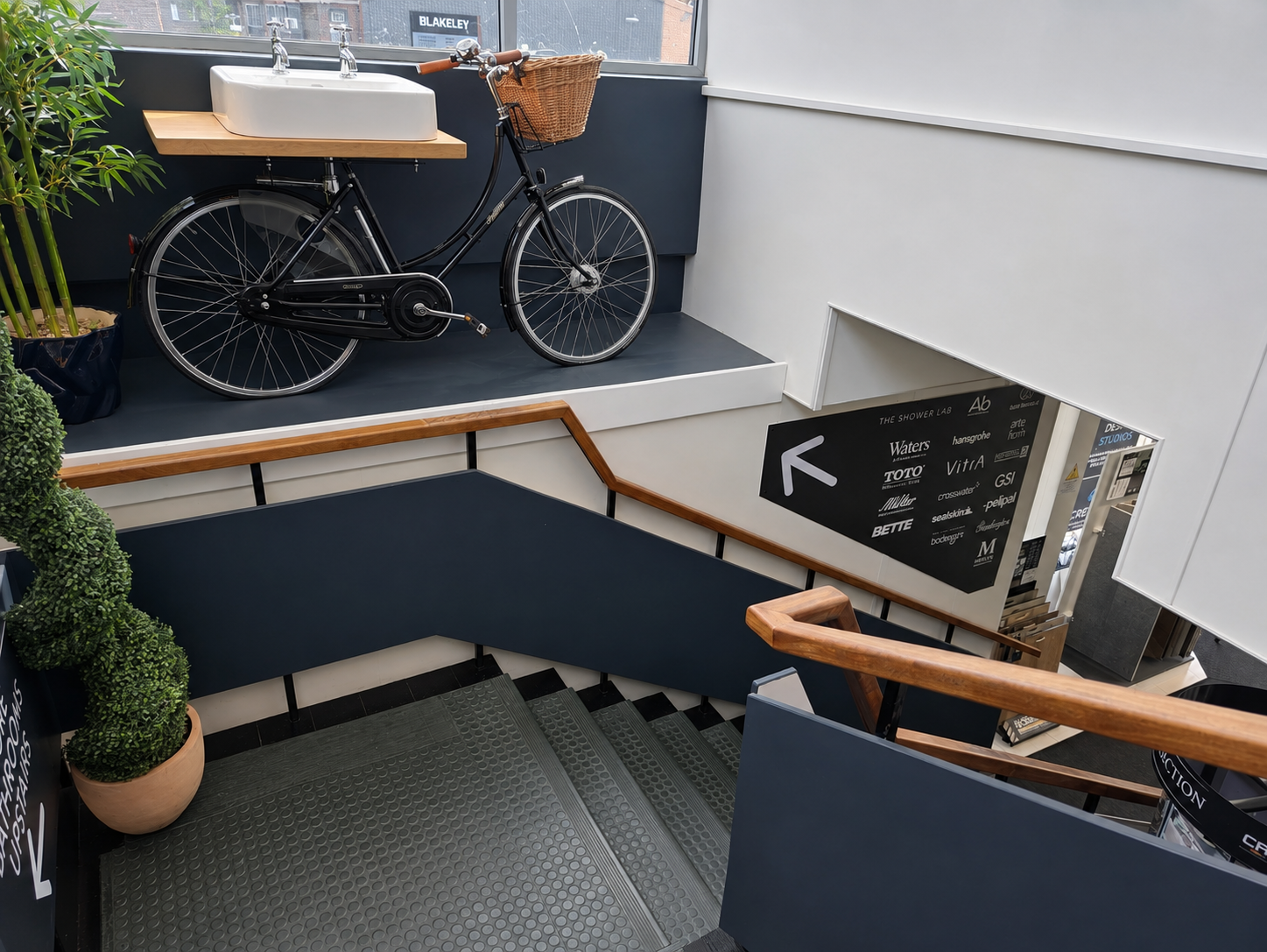 Interior view of a staircase with blue and wooden railings, a potted plant, and a staircase leading down. On the upper level, there is a black bicycle with a front basket parked near a window.