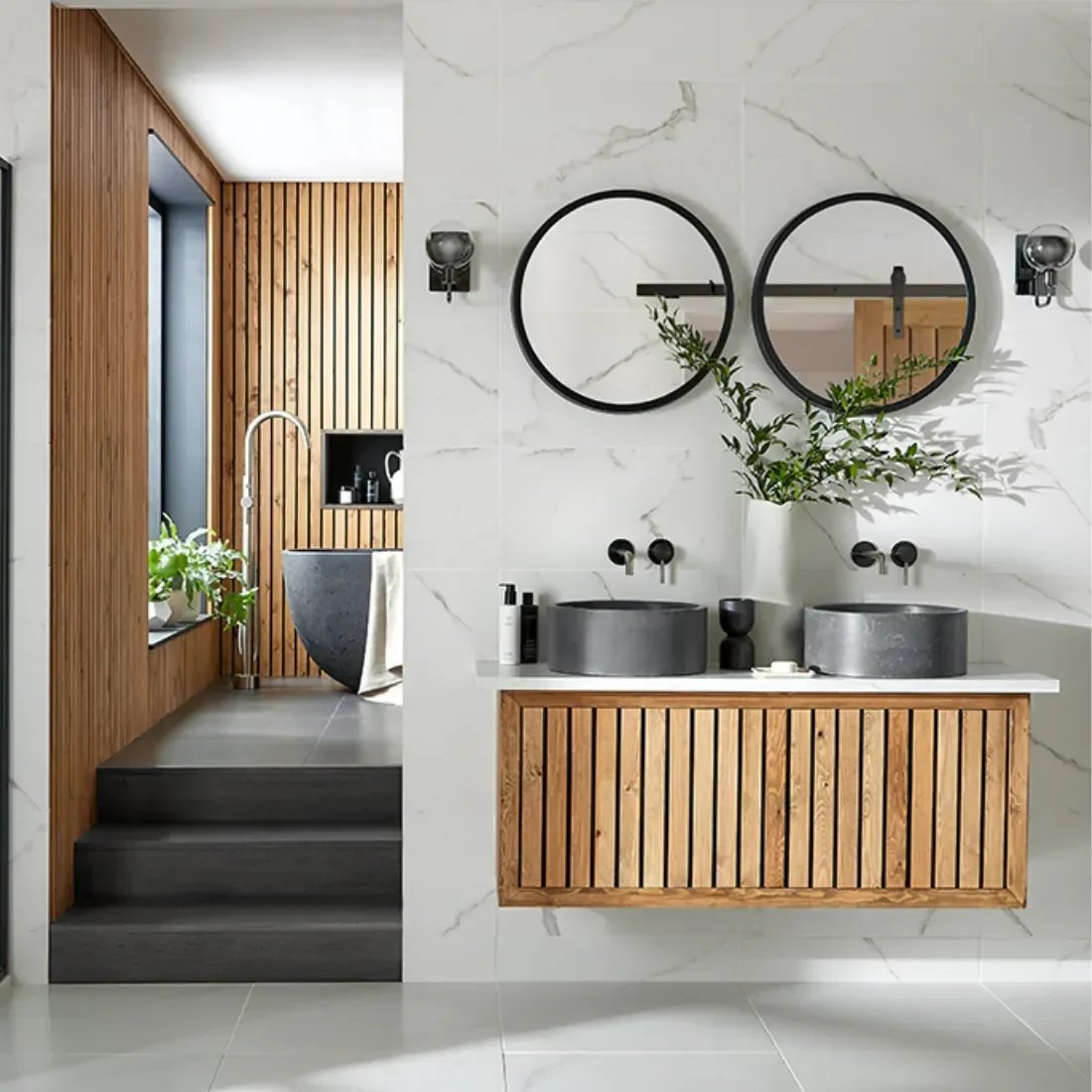 Modern bathroom with white marble wall, two round black-framed mirrors, and black vessel sinks on a wooden vanity. A wooden-accented wall with step leading to a separate bathing area with a freestanding bathtub. Items available in Island Bathrooms