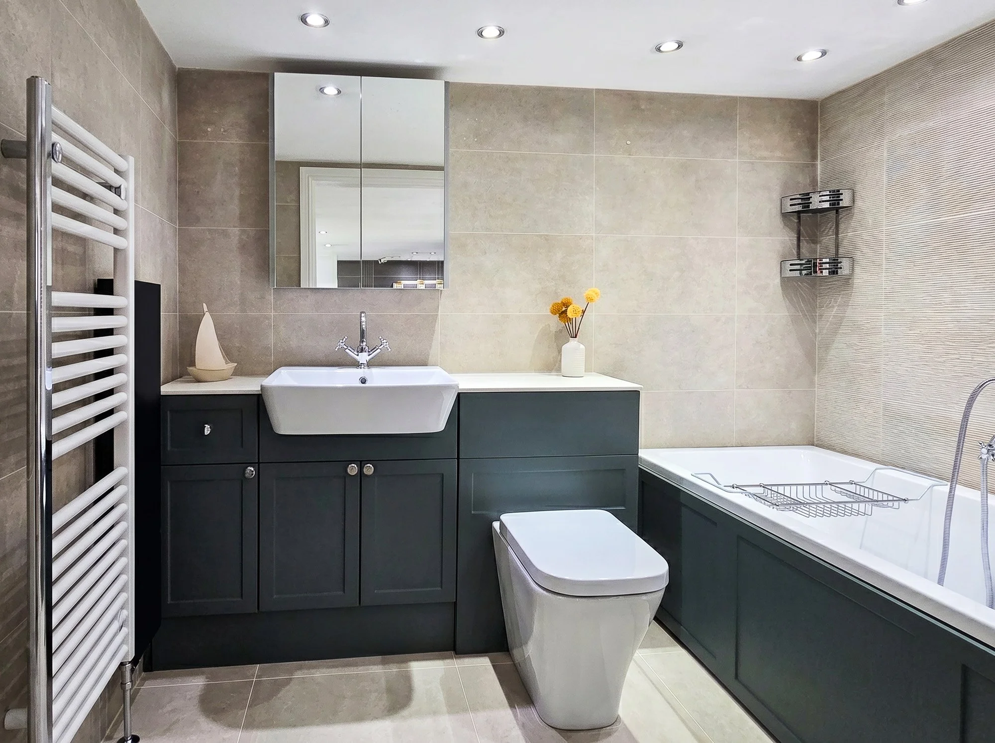 Modern bathroom with beige tiled walls, a white sink with storage cabinet beneath, a mirror cabinet above, a toilet, a heated towel rail, a vase with yellow flowers, and a bathtub with a drying rack inside.