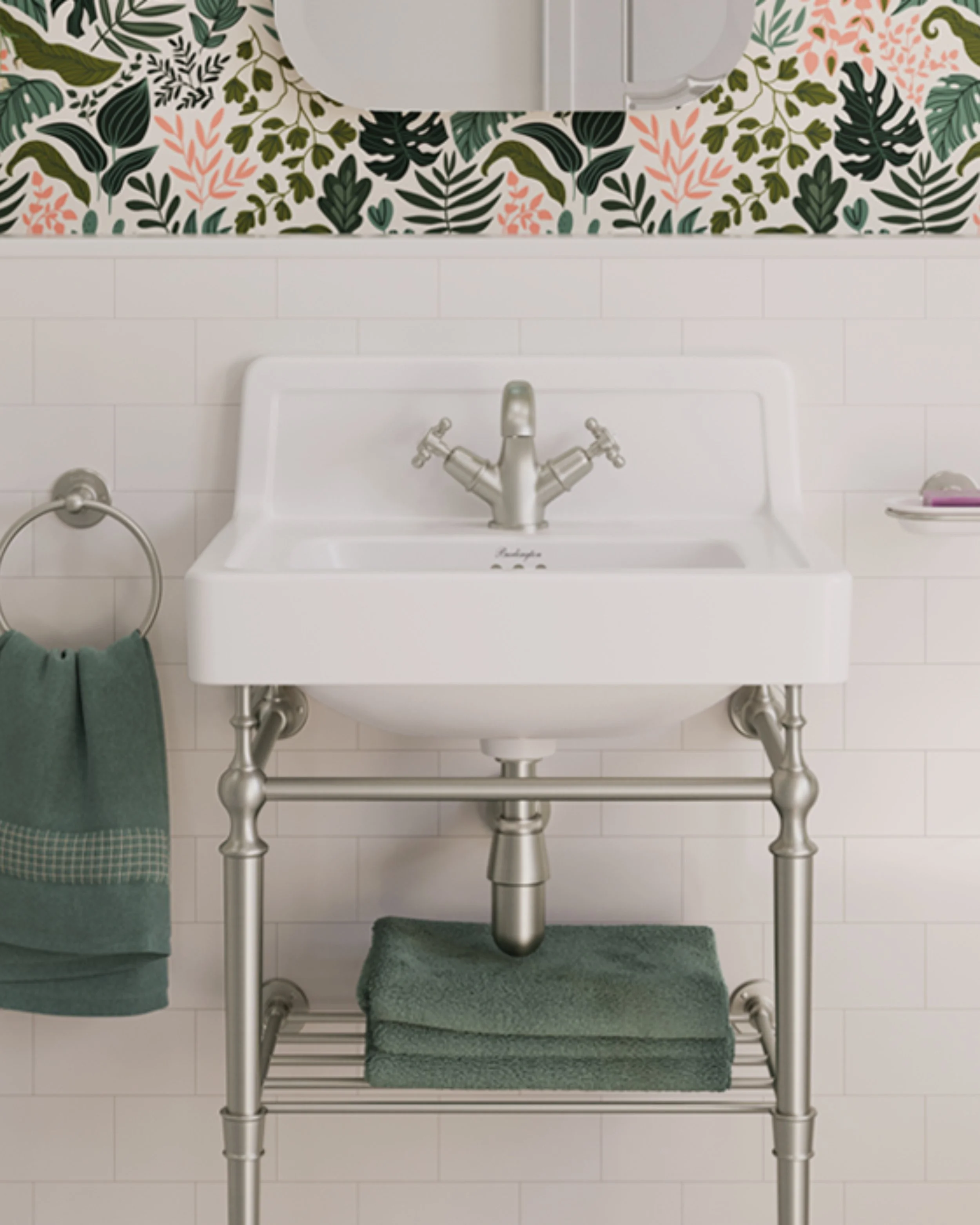 A white laundry sink with a silver faucet, positioned in front of a white tiled wall. A dark green towel hangs on a towel ring to the left. The sink is elevated on a metallic frame with a lower rack, on which a green towel is folded. Above the sink, 