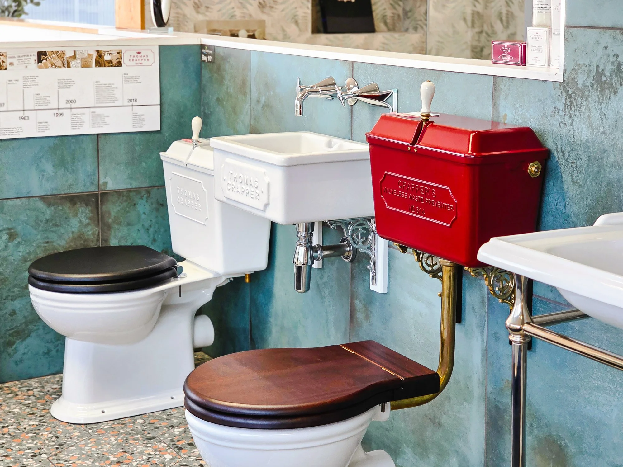 Restroom with vintage-style fixtures, including a red mailbox sink, a white ceramic sink, and a toilet with a black seat. The walls are tiled in teal, and there is a dark wood bench below the toilet. The floor is terrazzo.