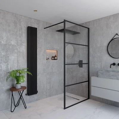 Modern bathroom with a glass shower enclosure, gray concrete walls, a wall-mounted white sink, a black circular mirror, a black vertical radiator, a small wooden side table with a green plant, and a minimalist niche in the wall for toiletries.