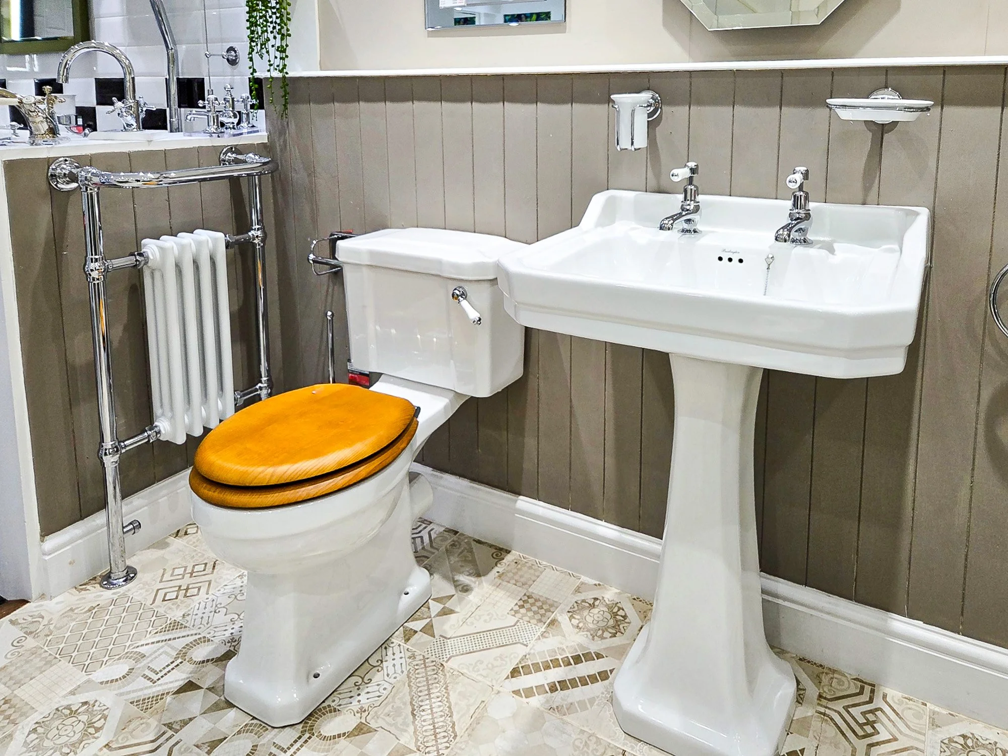 A vintage bathroom features a porcelain toilet with a wooden seat, a corner sink with two faucets, a wall-mounted radiator, and a mirror. The walls are partially covered in beige wood paneling with a tiled floor.