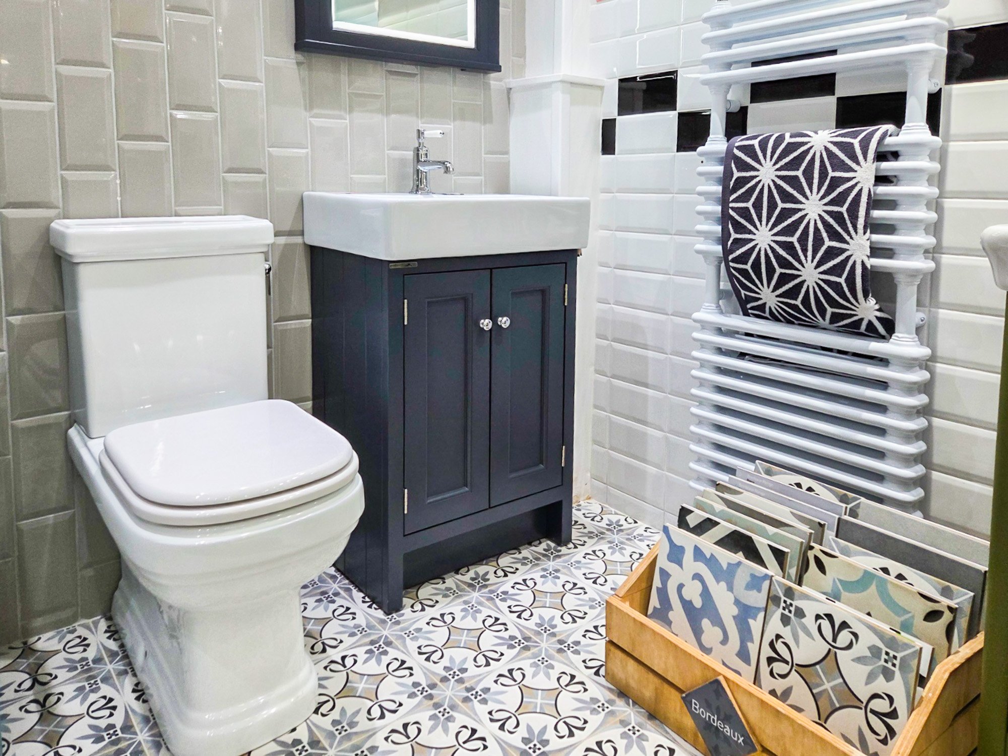 A bathroom featuring a toilet with a rectangular tank and rounded bowl, a small dark blue vanity with a white ceramic sink and modern chrome faucet, a medium-sized window, white wall tiles, and a black, white, and blue patterned ceramic floor. There is a white towel rack with a black and white patterned towel, a decorative tray with tile samples labeled 'Bordeaux', and a small bookcase with paint swatches and tiles.