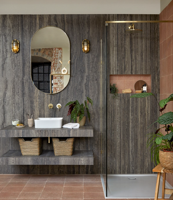 Modern bathroom with gray textured walls, oval mirror, gold fixtures, and a walk-in shower with dark wood paneling, pink tile, and a potted plant.