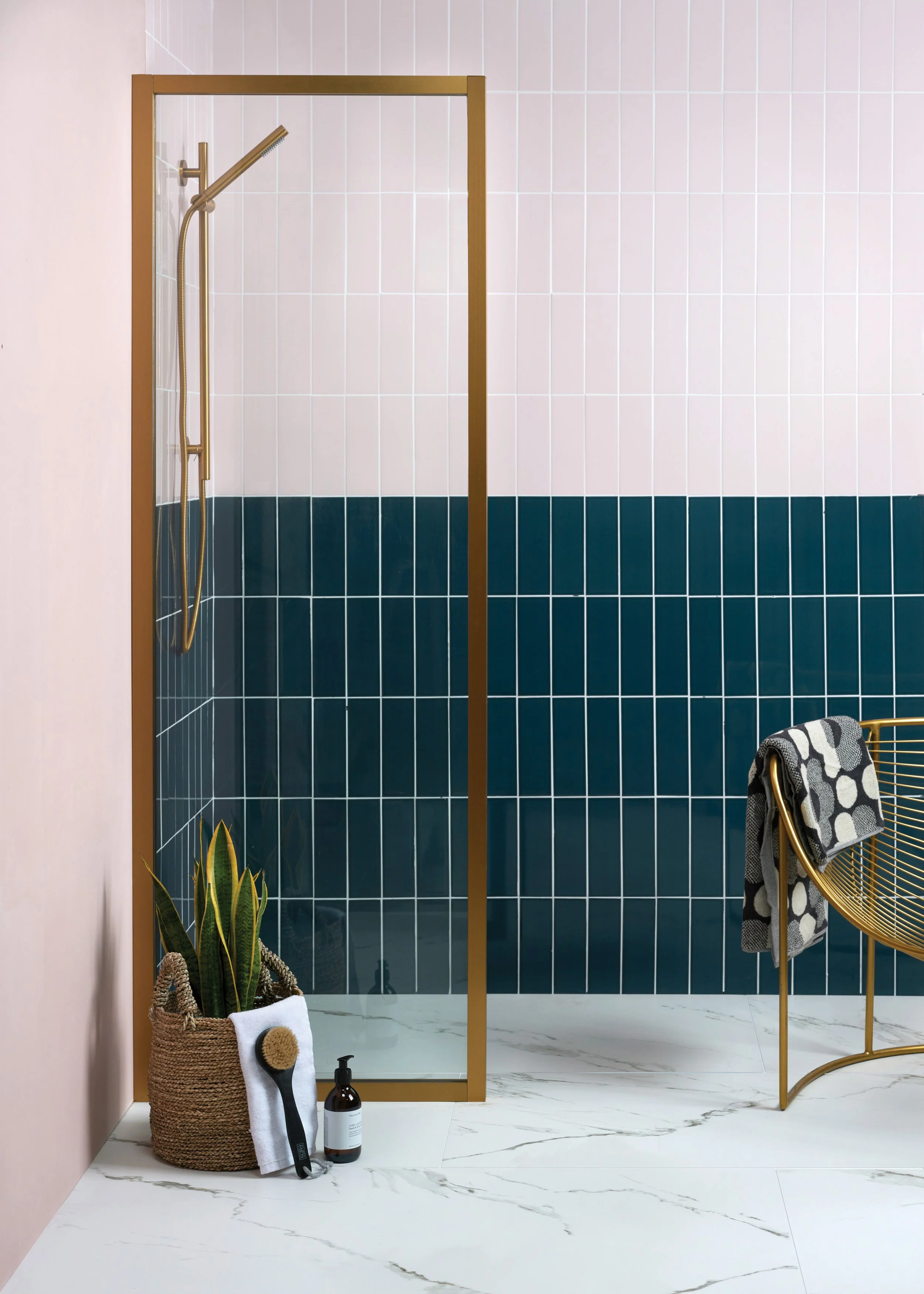 Modern bathroom with a glass shower enclosure, hanging towel, a potted snake plant, a black hairbrush, a soap dispenser, a gold chair with a black and white towel, and white marble flooring.