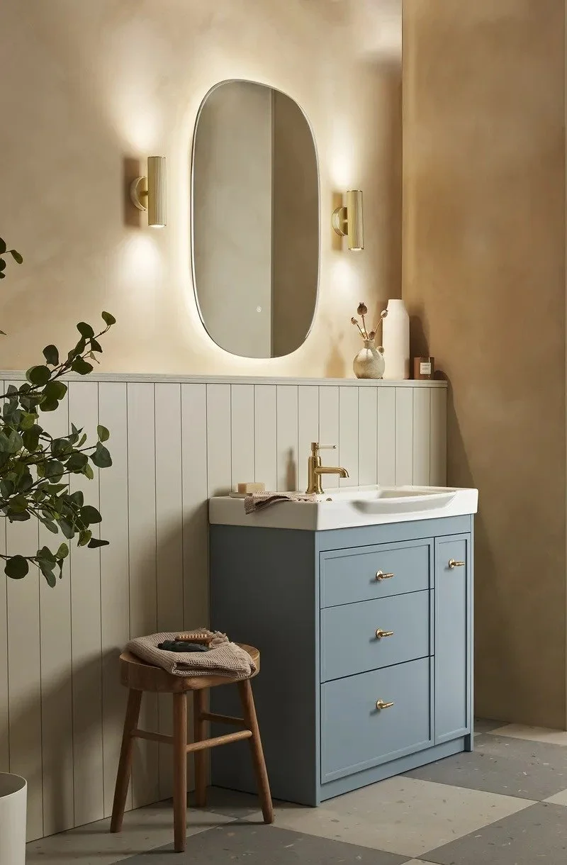 Bathroom with a blue vanity, oval mirror, wall-mounted lights, a small wooden stool, and decorative items including a vase with dried flowers.