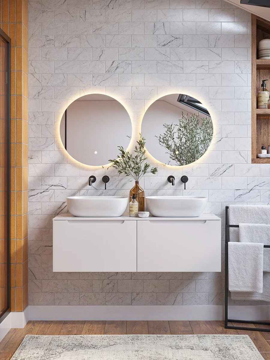 Modern bathroom with two white vessel sinks on a floating cabinet, two round backlit mirrors, a vase with green foliage, and towels hanging on a black metal towel rack.