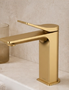 A gold-colored modern bathroom faucet on a white countertop with a beige wall in the background.