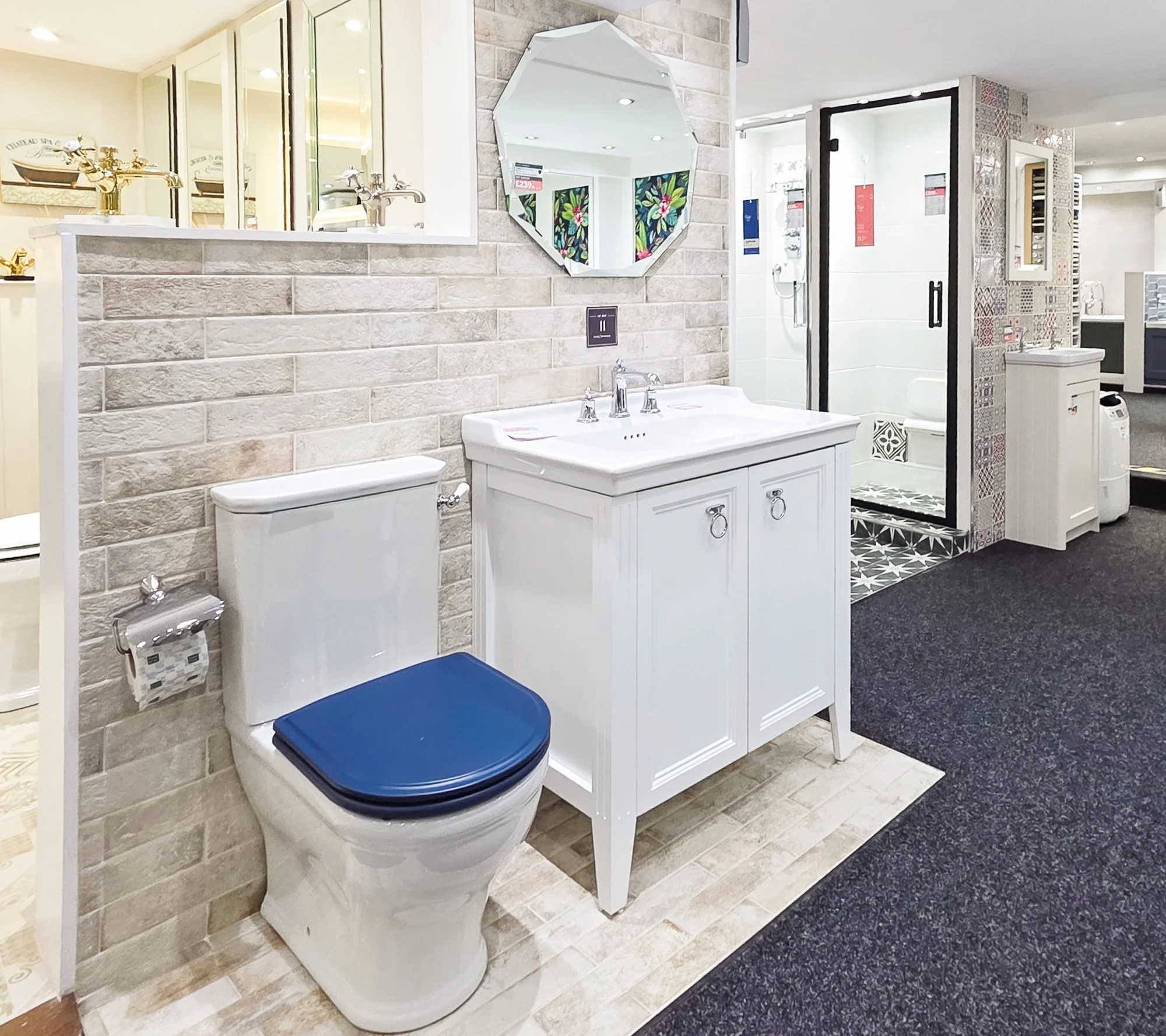 Bathroom display with toilet and vanity, featuring polished brass faucets, a mirror with a decorative frame, and shower area with patterned tiles and glass door, in a retail store setting.