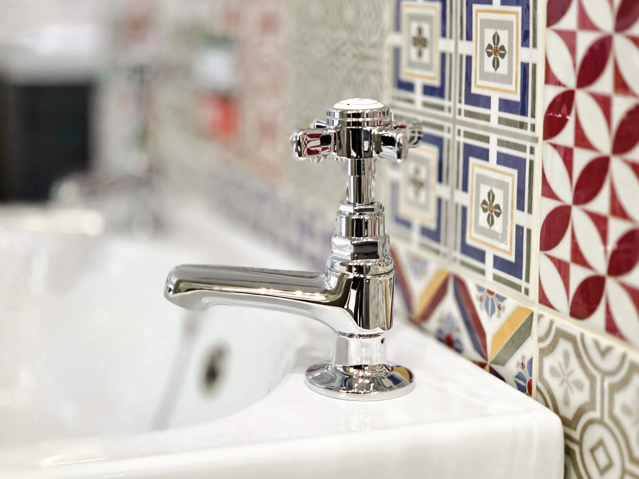 Close-up of a shiny chrome faucet on a white sink with decorative patterned tiles in the background.