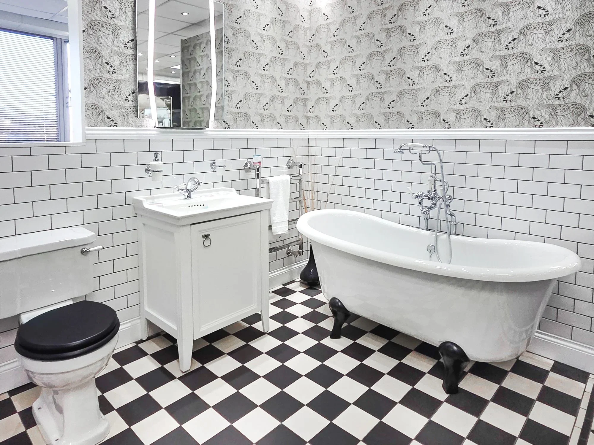 A vintage-style bathroom with white subway tile walls, a clawfoot bathtub, a small white sink, and a toilet with a black seat. The wall above the tiles features leopard print wallpaper. The floor has a black and white checkered pattern, and there is a window with blinds.