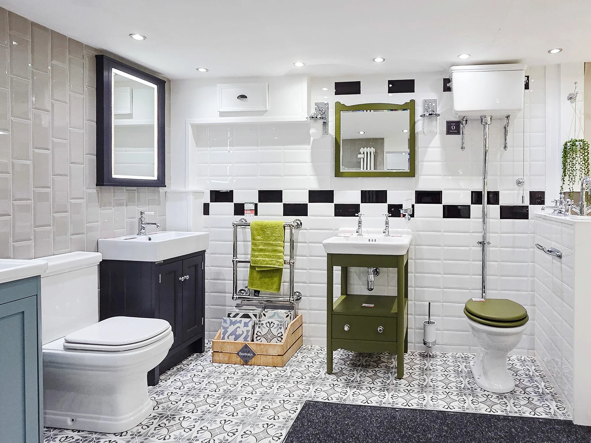 A modern bathroom with white tiled walls, patterned tile floor, and various fixtures including a toilet, sink, mirror, and shower, with colorful accents such as a green cabinet and matching towel.