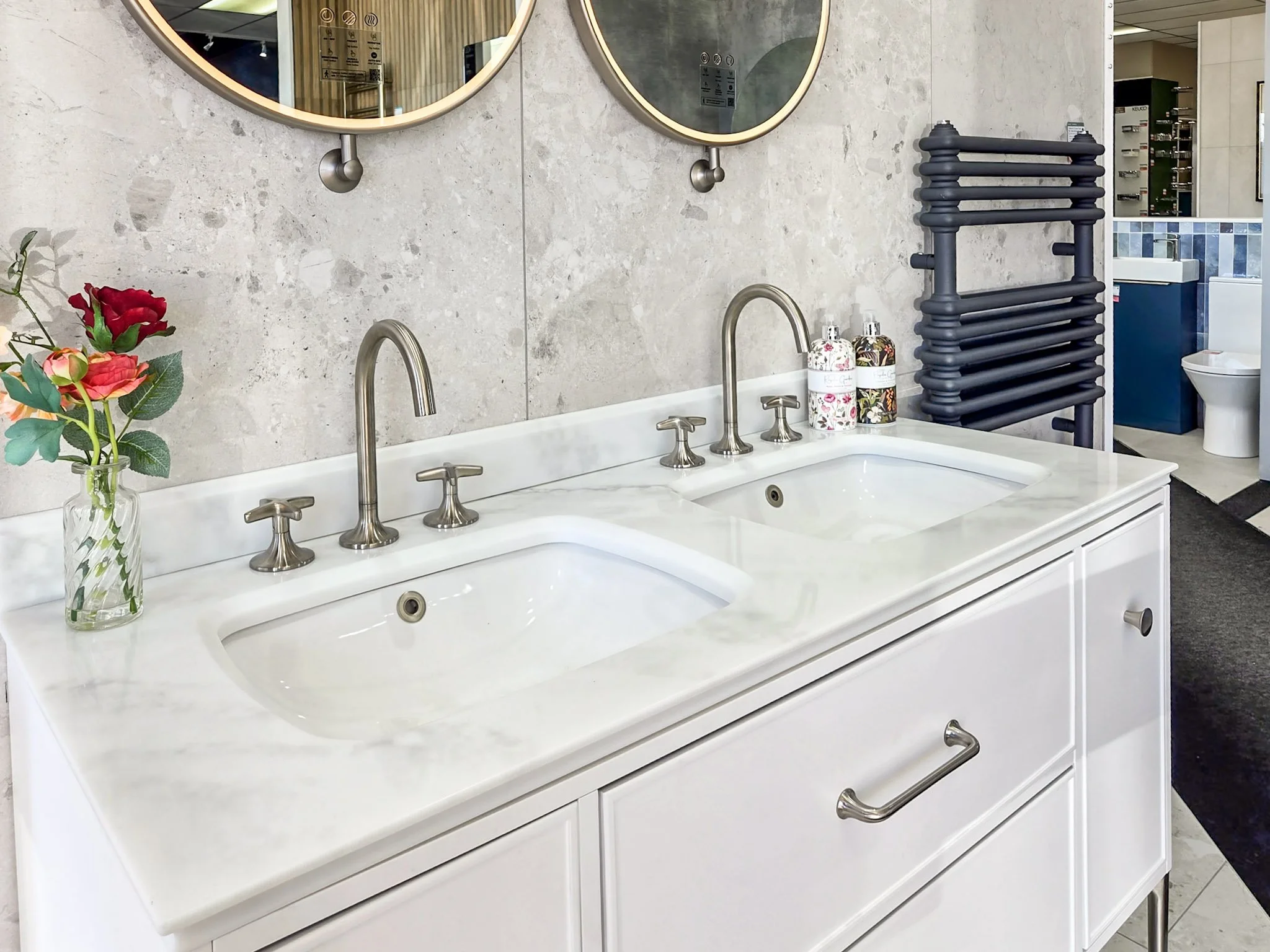 A white bathroom sink vanity with two sinks, each with a brushed nickel faucet. The vanity has a marble countertop and a single drawer with silver handles. To the left, there is a clear glass vase with red and pink flowers. To the right, there are two soap dispensers with floral patterns. Behind the vanity, there is a gray textured wall with two large round mirrors and black wall-mounted light fixtures. To the right of the vanity, there is a black heated towel rack and in the background, there is a dark towel hanging near a blue and white-tiled bathroom area with a white toilet.