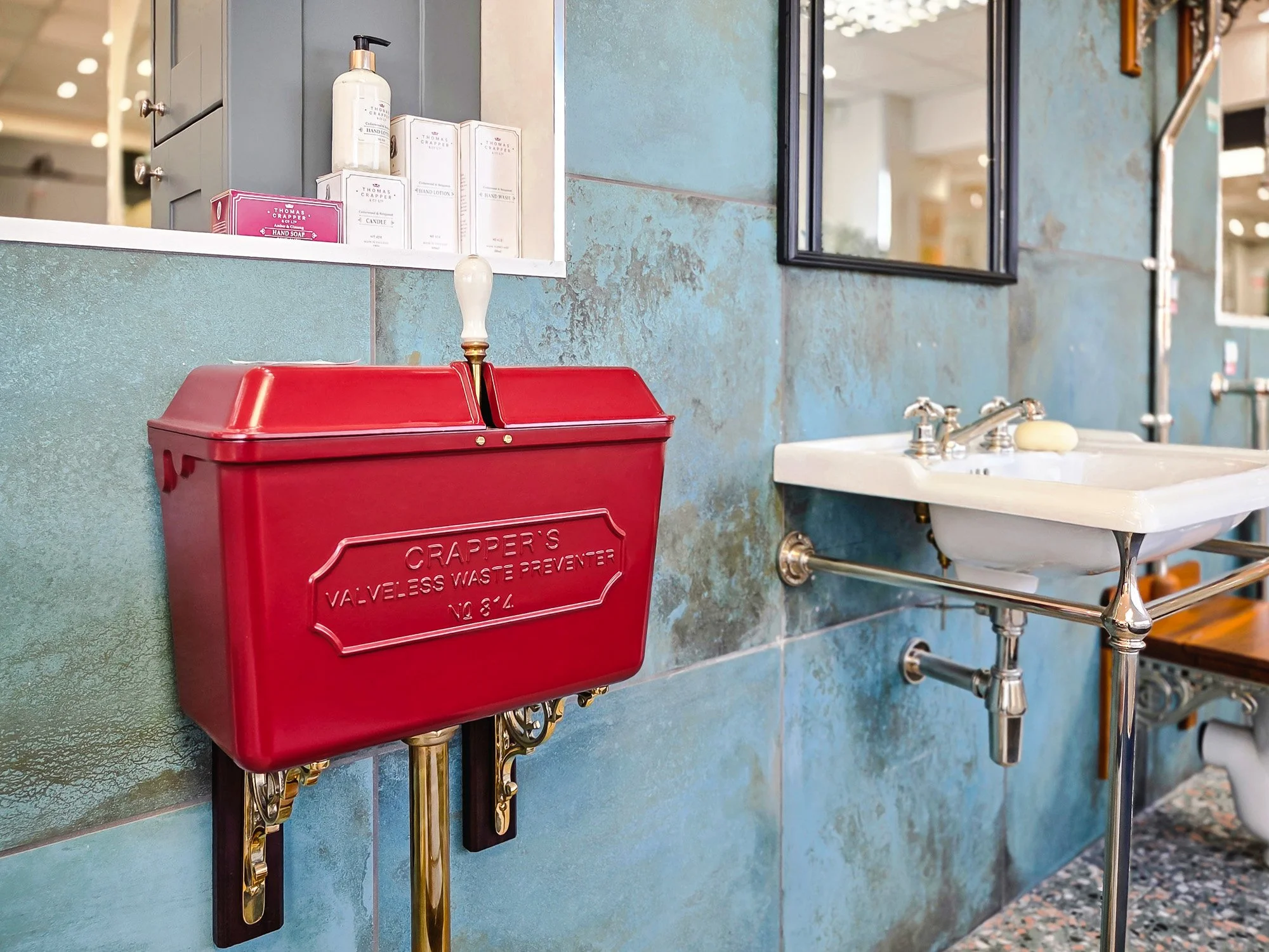 A red vintage valve-less waste water collector with gold accents, mounted on a green tiled wall near a white sink and mirror, with boxes of hand soap and lotion on a nearby ledge.