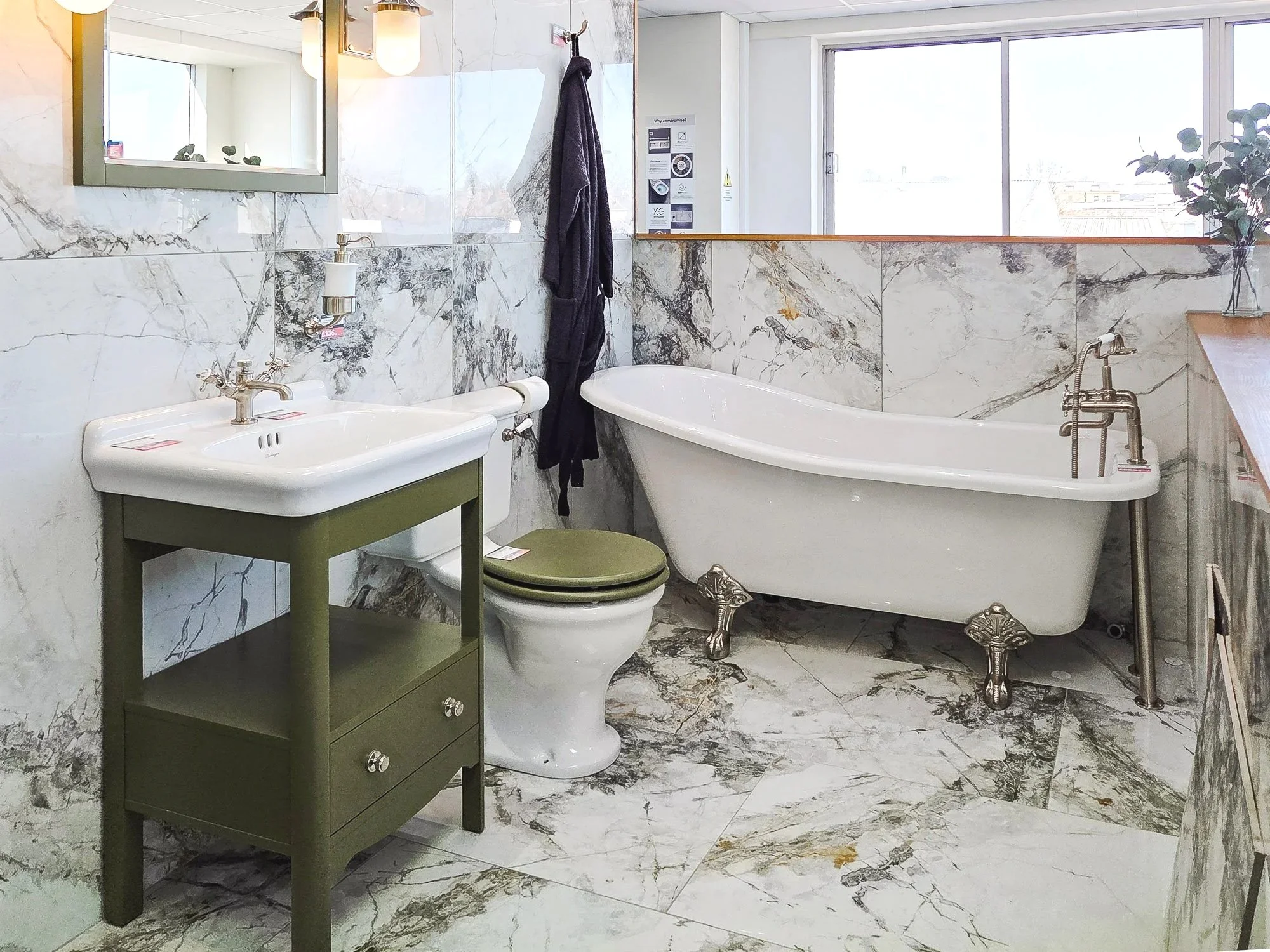 A luxurious bathroom with marble walls and floors, featuring a clawfoot bathtub, a small green vanity with drawers, a toilet, a wall mirror, and a window that lets in natural light. Decor includes a dark towel hanging on a hook, a clear vase with green foliage on a counter, and wall-mounted lighting fixtures.