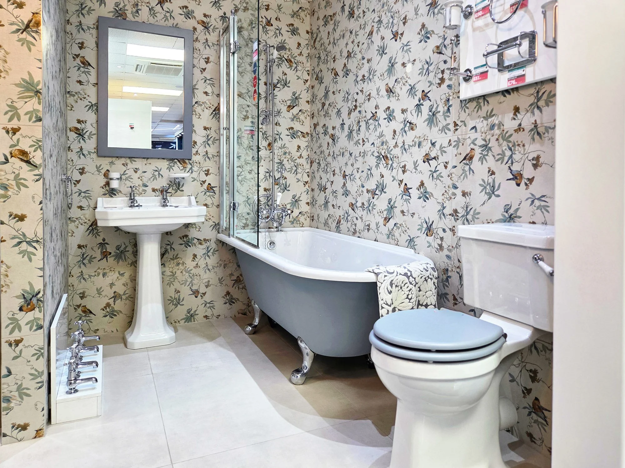 A bathroom with bird and foliage patterned wallpaper, a vintage clawfoot bathtub, a white pedestal sink, a mirror, and a toilet. There is a shower with glass doors and chrome fixtures.