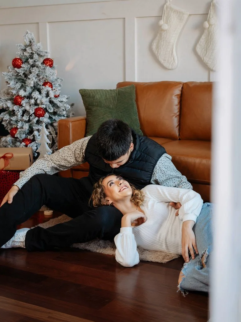 A woman lying on the floor and smiling at a boy on top of her, in a cozy living room decorated for Christmas.