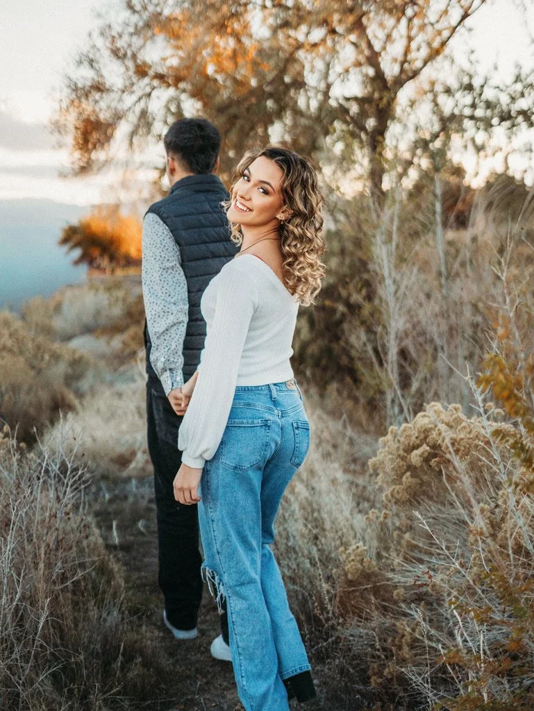 A woman with curly hair smiling at the camera, standing outdoors on a nature trail near a man with short dark hair, outdoors with autumn trees in the background.